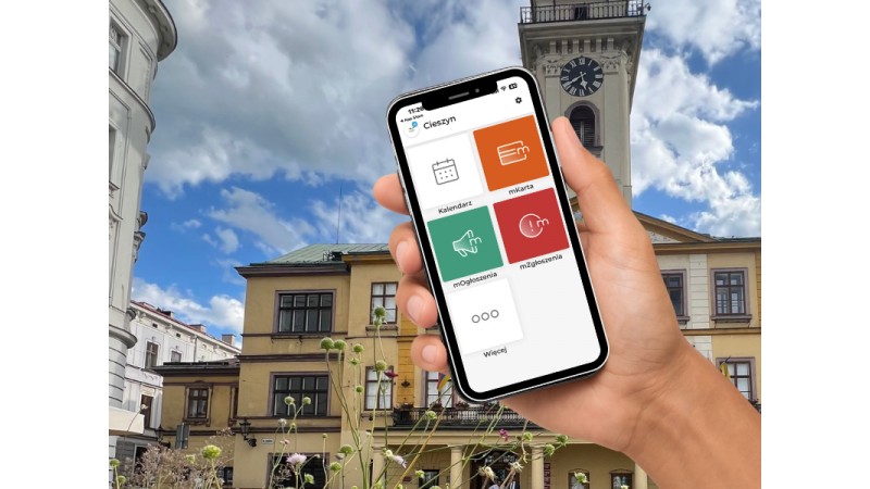 Na zdjęciu widać dłoń trzymającą smartfon na tle cieszyńskiego Ratusza. W telefonie jest otwarta aplikacja Cieszyn z kafelkami: Kalendarz, mKarta, Ogłoszenia, Zgłoszenia i Więcej. W tle znajduje się rynek z kamienicami oraz wieża z zegarem